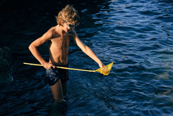 Criança em calções de banho e óculos de mergulho com rede de colher em pé no mar ondulado em raio de sol - Foto, Imagem