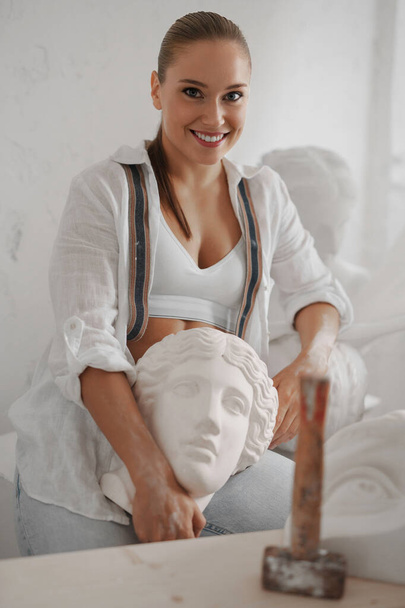 A talented female sculptor, dressed in a white shirt and jeans with suspenders, poses serenely in her workshop, showcasing her craftsmanship with a vintage Greek bust - Photo, Image