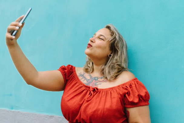 Boční pohled na mladé plus velikost žena v ležérní oblečení přičemž selfie v kulaté tlamě pózovat s chytrým telefonem, zatímco stojí na modré zdi pozadí venku - Fotografie, Obrázek