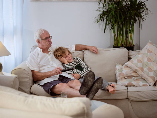 Volledig lichaam van verrukt kleinzoon kijken film met senior man terwijl rusten op de bank in lichte woonkamer thuis - Foto, afbeelding