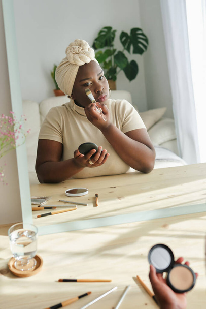 Reflejado en espejo Afro-Americano hembra en turbante aplicando polvo en la cara usando cepillo en casa - Foto, imagen