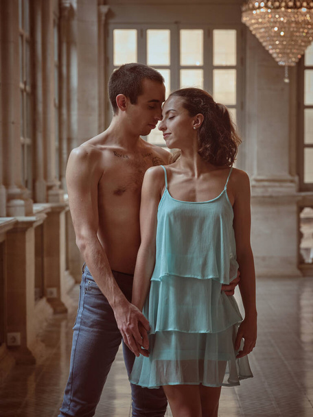 Shirtless man in denim and girlfriend in dress looking at each other and holding hands while standing together in hall - Photo, Image