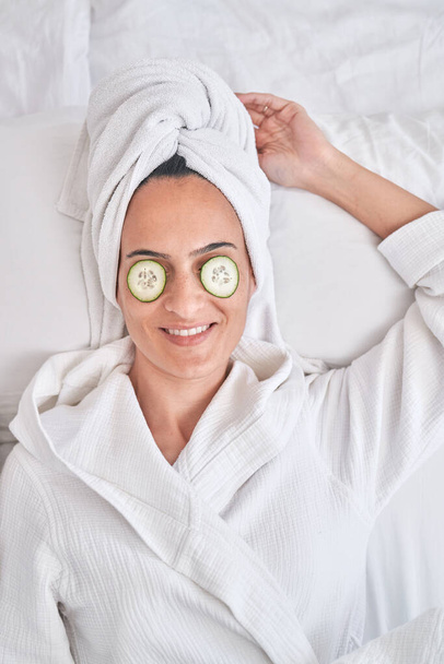From above of relaxed female in bathrobe and towel with cucumber slices on eyes resting on bed during skincare procedure - Photo, Image