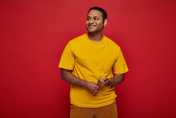 ihana intialainen mies kirkkaissa rento vaatteissa katselee pois ja hymyilee punaisella taustalla studiossa - Valokuva, kuva