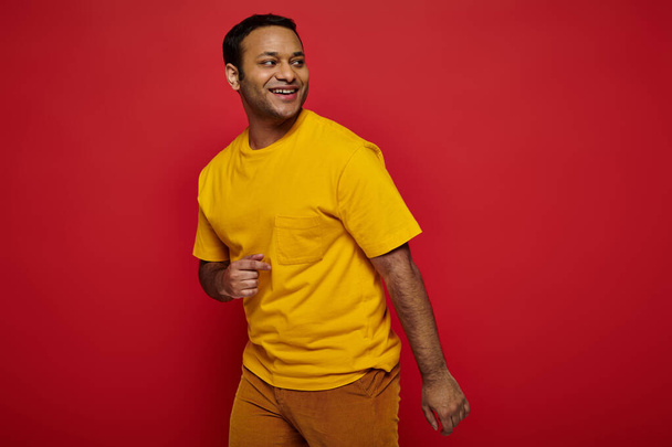 stralende indiaanse man in heldere casual kleding weg te kijken en te glimlachen op rode achtergrond, optimist - Foto, afbeelding