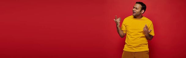 эмоциональный индиец в яркой повседневной одежде жестом, объясняя что-то на красный, баннер - Фото, изображение