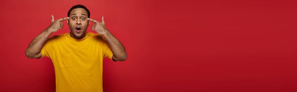 умопомрачительный, шокированный индиец с пальцами возле головы, смотрящий на камеру на красном фоне, баннер - Фото, изображение