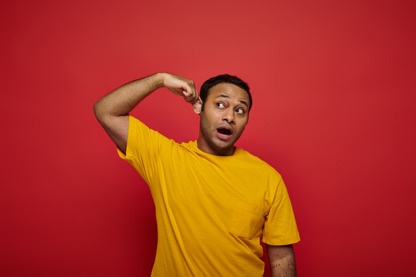 curious indian man in bright casual clothes looking away and touching ear on red background - Photo, Image