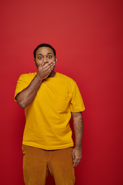 shocked indian man in bright clothes covering mouth and looking at camera on red background - Photo, Image