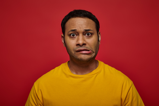 worried indian man in bright clothes looking at camera and grimacing on red background, stroke - Foto, Imagem