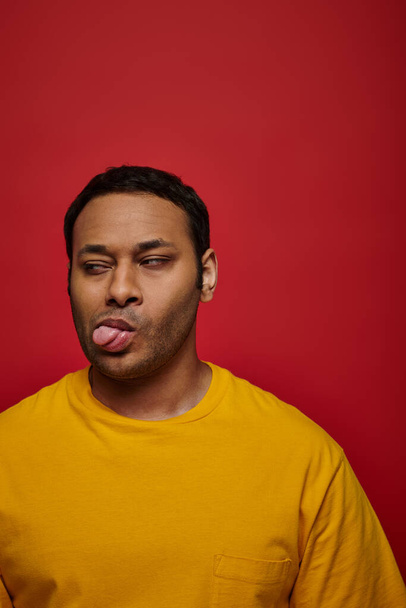 face expression, displeased indian man in yellow clothes sticking out tongue on red backdrop - 写真・画像