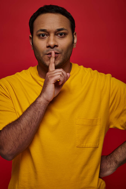 uomo indiano in abiti gialli che mostra il segno del silenzio e guardando la fotocamera sullo sfondo rosso, segreto - Foto, immagini