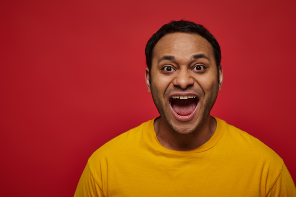 emoción positiva, hombre indio excitado en camiseta amarilla con la boca abierta sobre fondo rojo - Foto, Imagen