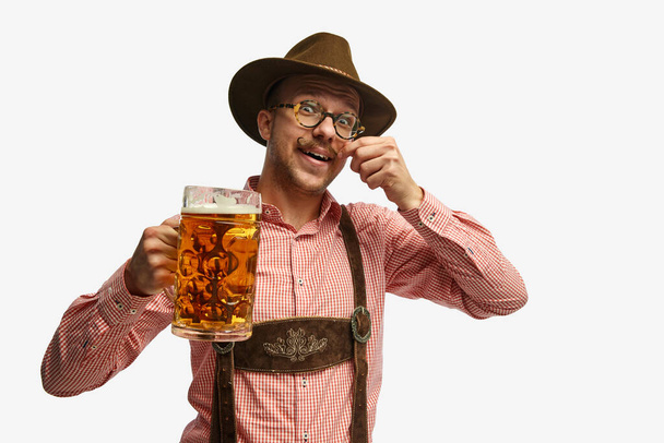 Ça a l'air heureux. Portrait de jeune homme séduisant au chapeau, portant des vêtements bavarois folkloriques, tenant une tasse de bière isolée fond blanc. Alcool, traditions, vacances, festival et concept publicitaire. - Photo, image