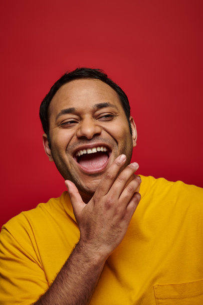 émotion positive, excité homme indien en t-shirt jaune riant avec bouche ouverte sur fond rouge - Photo, image