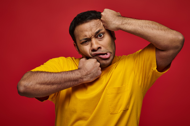expressão facial, homem indiano confuso em camiseta amarela perfurando-se no rosto em pano de fundo vermelho - Foto, Imagem