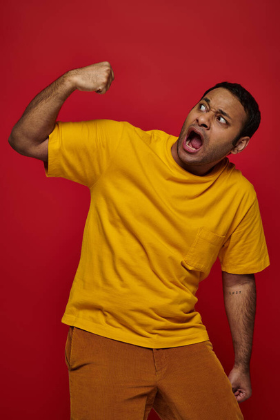 face expression, scared indian man in yellow t-shirt punching himself into face on red background - Photo, Image