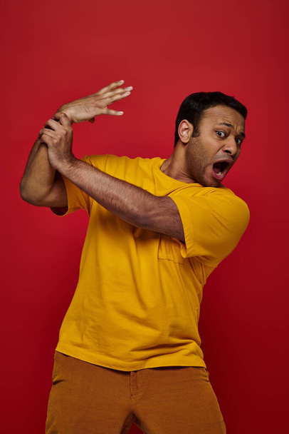Yüz ifadesi, sarı tişörtlü şok olmuş Hintli adam kırmızı arka planda elini kaybediyor. - Fotoğraf, Görsel