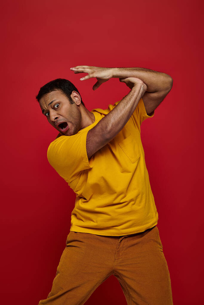 face expression, hand on scared indian man in yellow t-shirt attacking him on red background - Photo, Image