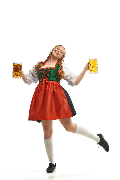 Kelnerka. Pełnowymiarowy portret tańczącej uroczej młodej kobiety noszącej ludowy niemiecki dirndl z dwoma kubkami piwa na białym tle. Koncepcja Oktoberfest, tradycje, napoje i jedzenie. Kopiuj miejsce na reklamę - Zdjęcie, obraz