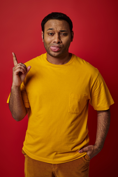 gezichtsuitdrukking, emotionele man steken uit tong en wijzen op rode achtergrond, grimas - Foto, afbeelding
