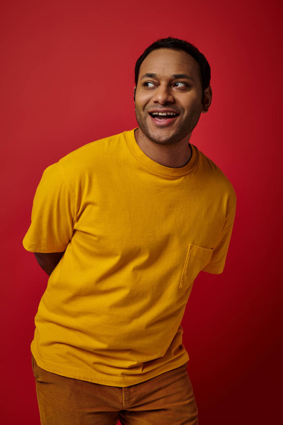 hombre indio alegre en camiseta amarilla mirando hacia otro lado y sonriendo sobre el fondo rojo, expresión de la cara - Foto, imagen