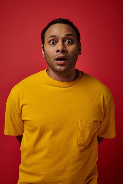 erstaunt indischer Mann in gelbem T-Shirt blickt in die Kamera auf rotem Hintergrund, Gesichtsausdruck - Foto, Bild