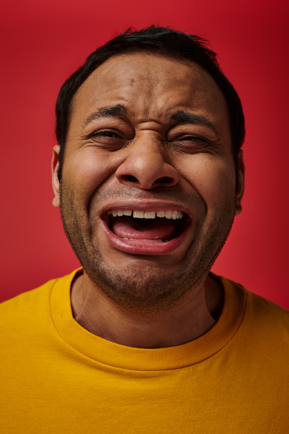 expresión de la cara, hombre indio emocional en camiseta amarilla llorando fuerte sobre fondo rojo, boca abierta - Foto, Imagen