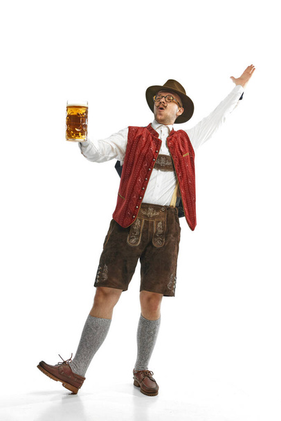 Τραγουδώντας χαρούμενα. Νεαρός συναισθηματικός μουστακαλής με καπέλο, φοράει βαυαρικά ή γερμανικά ρούχα, κρατάει κούπα μπύρας. Έννοια του αλκοόλ, παραδόσεις, διακοπές, γιορτή. Αντιγραφή χώρου για διαφήμιση - Φωτογραφία, εικόνα