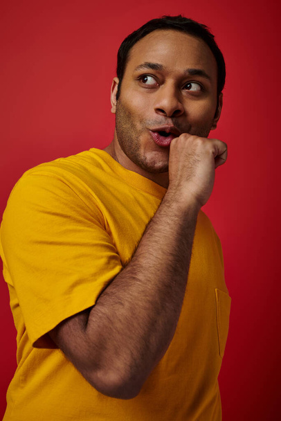 výraz obličeje, indický muž ve žlutém tričku zobrazující reakci při pohledu jinam na červené pozadí - Fotografie, Obrázek