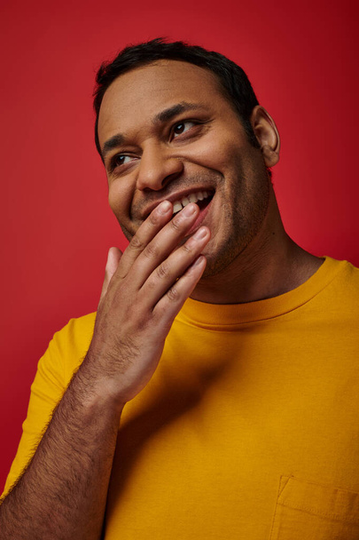 shy indian man in yellow t-shirt smiling and covering mouth with hand on red background in studio - Photo, Image