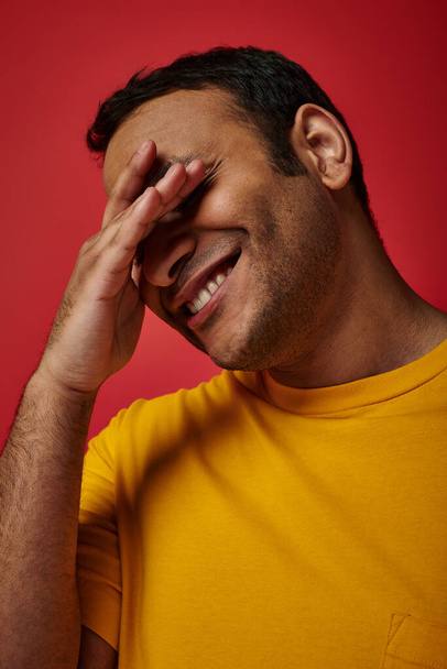 shy indian man in yellow t-shirt smiling and covering eyes with hand on red background in studio - Photo, Image