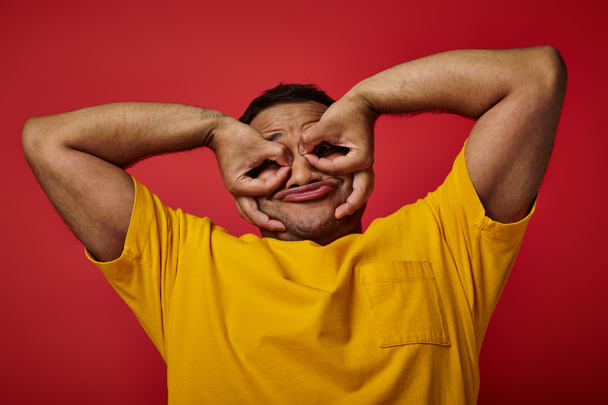 funny indian man in yellow t-shirt grimacing and gesturing on red backdrop, expressive face - Photo, Image