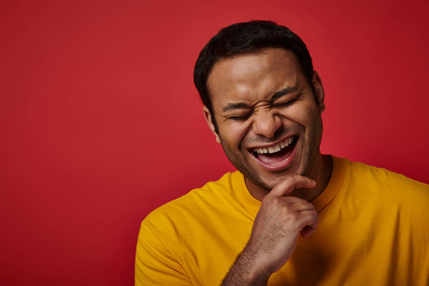 gioioso uomo indiano in t-shirt gialla sorridente ad occhi chiusi su sfondo rosso in studio, ritratto - Foto, immagini
