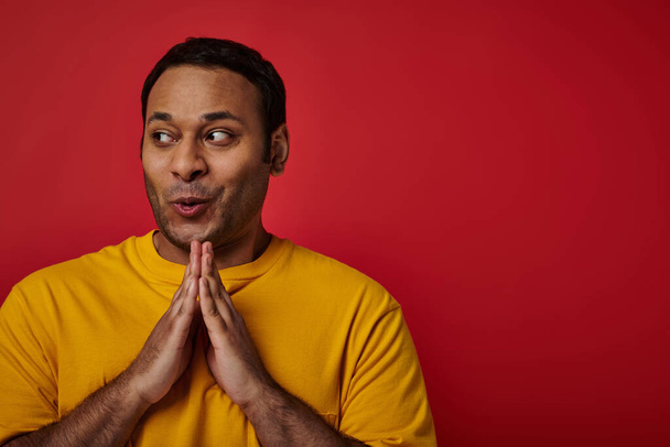 astuto hombre indio en camiseta amarilla agarrándose de las manos y planeando algo sobre fondo rojo, cara astuta - Foto, Imagen