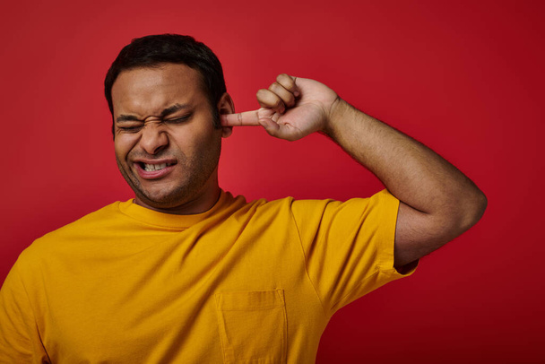индиец в жёлтой футболке закрывает глаза и ковыряется пальцем в ухе на красном фоне, гримаса - Фото, изображение