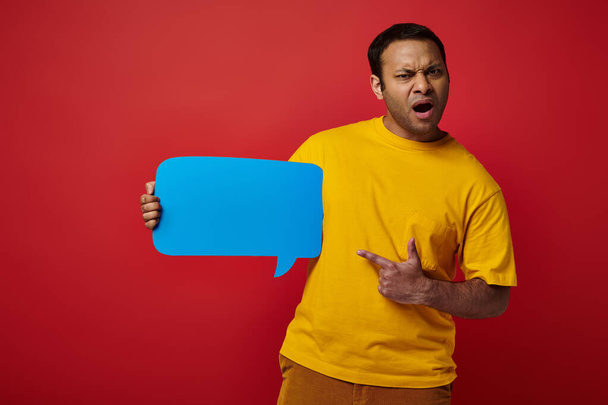 hombre sorprendido en camiseta amarilla apuntando a la burbuja del habla en blanco sobre el fondo rojo, disgustado - Foto, Imagen
