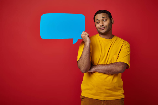 skeptical indian man in yellow t-shirt holding blank speech bubble on red background - Photo, Image