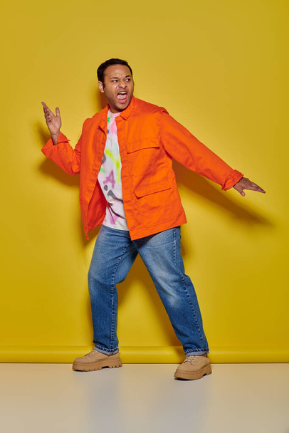 homem índio emocional em casaco laranja e jeans gesticulando e gritando em pano de fundo amarelo - Foto, Imagem