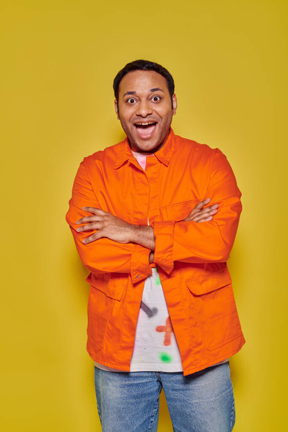 portrait of amazed indian man in orange jacket standing with folded arms on yellow backdrop - Photo, Image