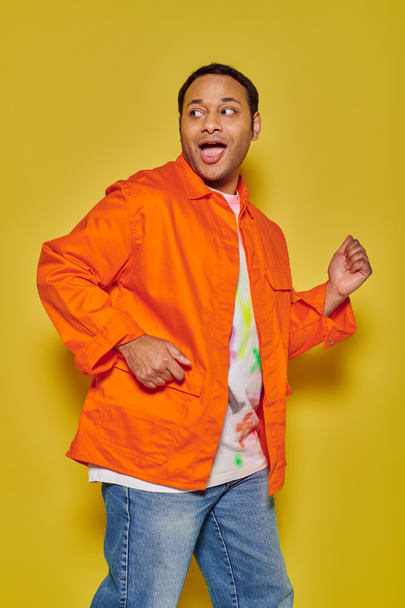 portrait of amazed indian man in orange jacket running away and looking back on yellow backdrop - Foto, afbeelding