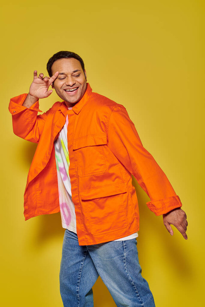 portrait of happy indian man in orange jacket and denim jacket dancing on yellow background - Photo, Image