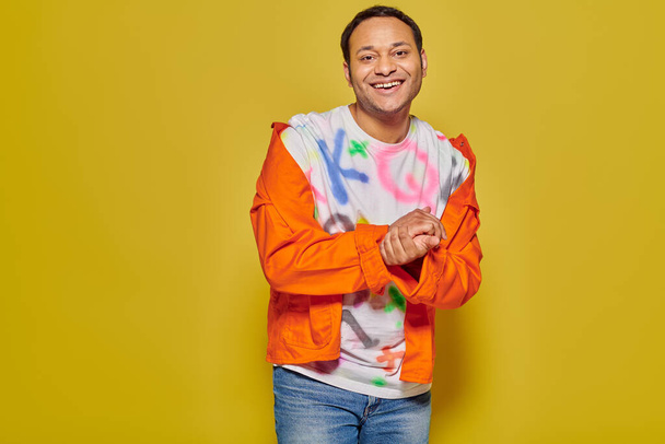 happy indian man in orange jacket and diy t-shirt smiling while looking at camera on yellow backdrop - Photo, Image