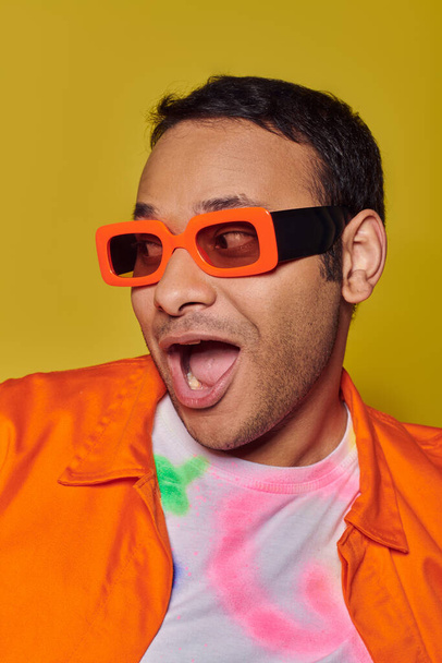 concepto de expresión personal, hombre indio excitado en gafas de sol naranjas sonriendo en el fondo amarillo - Foto, imagen
