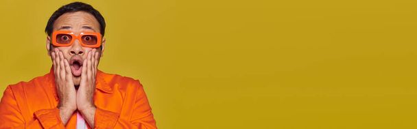 uomo indiano scioccato in occhiali da sole arancioni toccare il viso e dire wow su sfondo giallo, banner - Foto, immagini