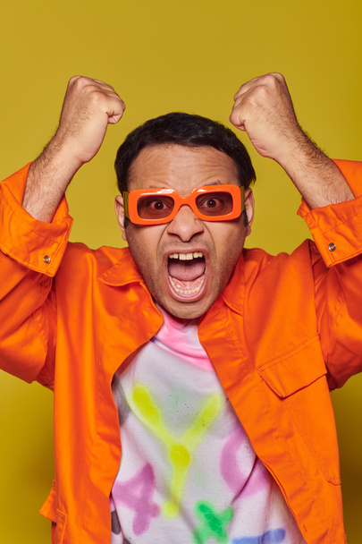 hombre indio enojado en gafas de sol naranjas mirando a la cámara y haciendo gestos sobre fondo amarillo - Foto, Imagen