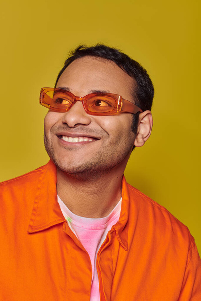 Turuncu güneş gözlüklü mutlu Hintli adam sarı arka planda gülümserken başka tarafa bakıyor. - Fotoğraf, Görsel