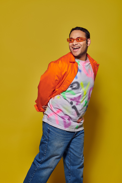 amazed indian man in sunglasses and vibrant attire looking away and smiling on yellow background - Photo, Image