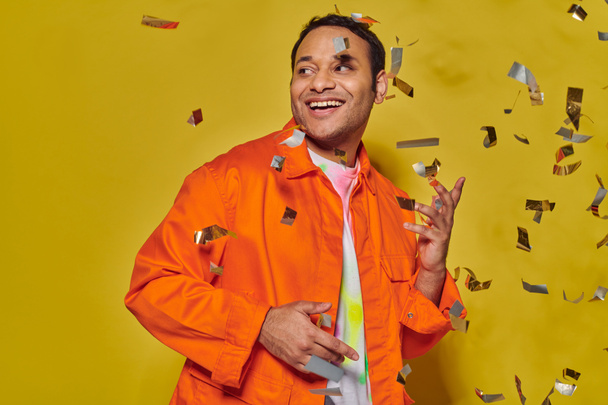hombre indio feliz en chaqueta naranja brillante sonriendo cerca de caer confeti sobre fondo amarillo, fiesta - Foto, imagen