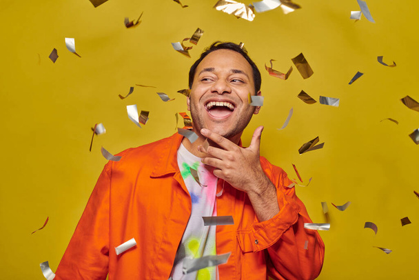 joyous indian man in bright orange jacket smiling near falling confetti on yellow backdrop, party - Photo, Image